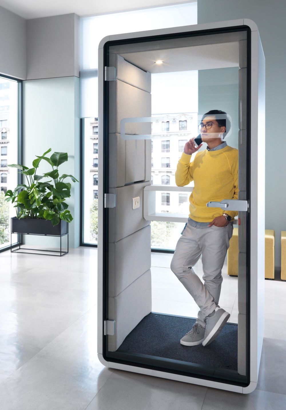 Person in gelbem Pullover telefoniert in schallisolierter Telefonzelle im modernen Büro mit großen Fenstern.