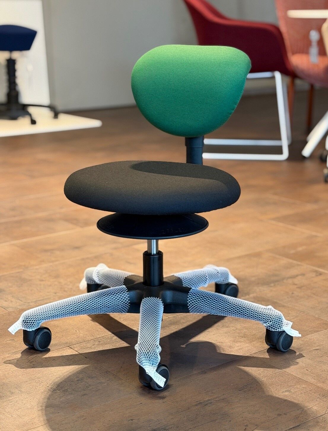 Ergonomischer Bürostuhl mit grünem Rücken und schwarzem Sitz auf einem Holzboden im Showroom ausgestellt.