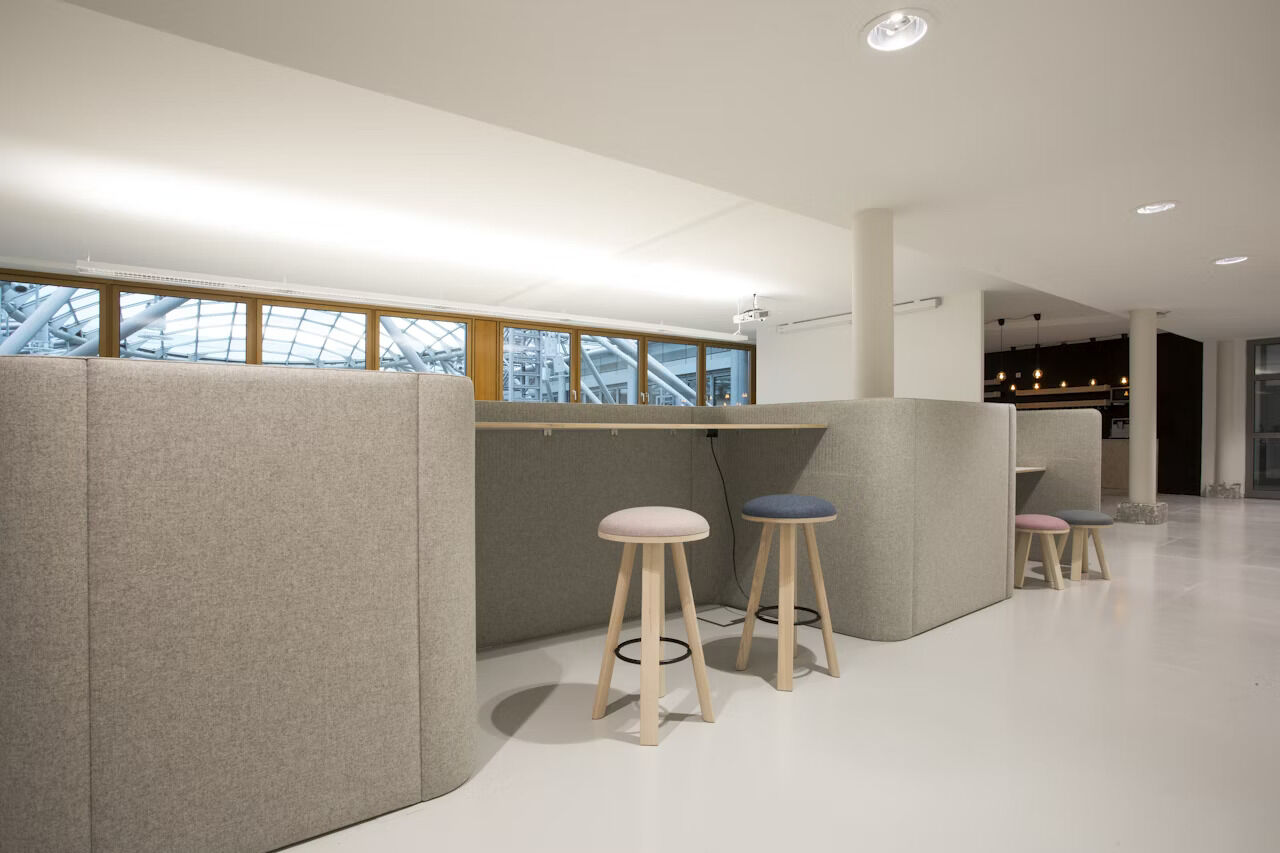 Moderne Büroeinrichtung mit hohen Tischen und gepolsterten Hockern, in hellem, offenem Raum angeordnet.