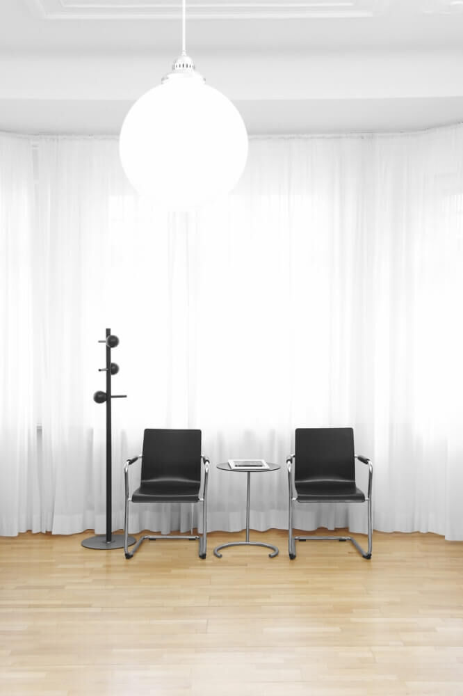 Zwei schwarze Bürostühle mit Beistelltisch und Garderobenständer vor weißen Vorhängen in einem minimalistischen Raum.