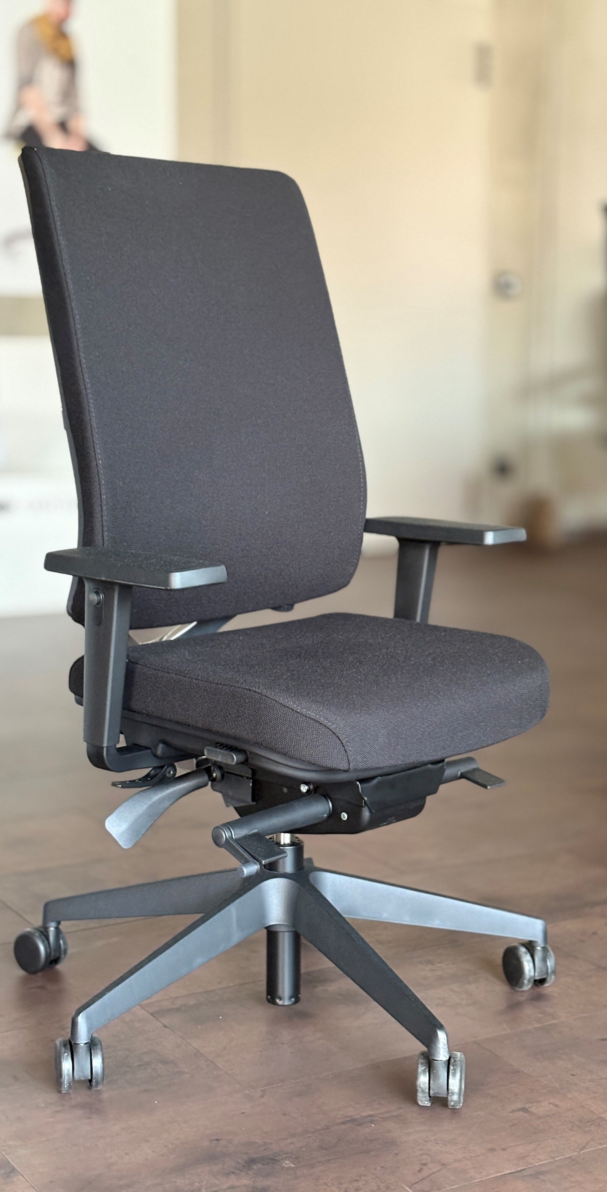 Ergonomischer Bürostuhl mit schwarzem Stoffbezug, verstellbaren Armlehnen und fünf Rollen auf Holzboden.