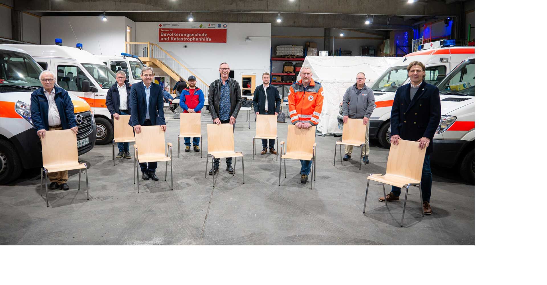 Gruppe von Menschen steht hinter Holzstühlen in einer Garage mit Rettungswagen im Hintergrund.