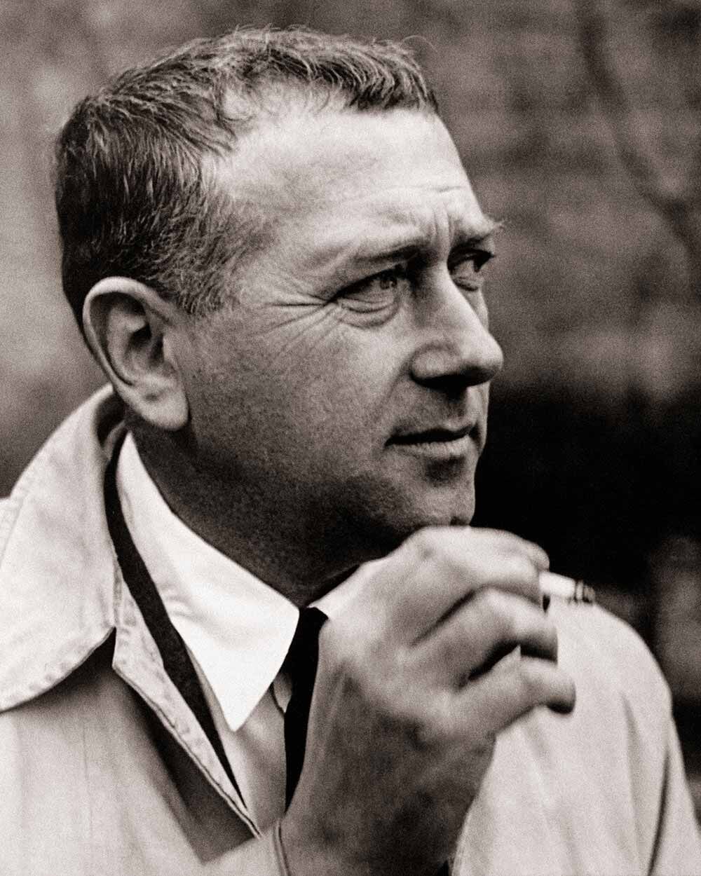 Ein Mann im Profil mit kurzer Frisur, trägt einen Trenchcoat, hält eine Zigarette in der Hand.