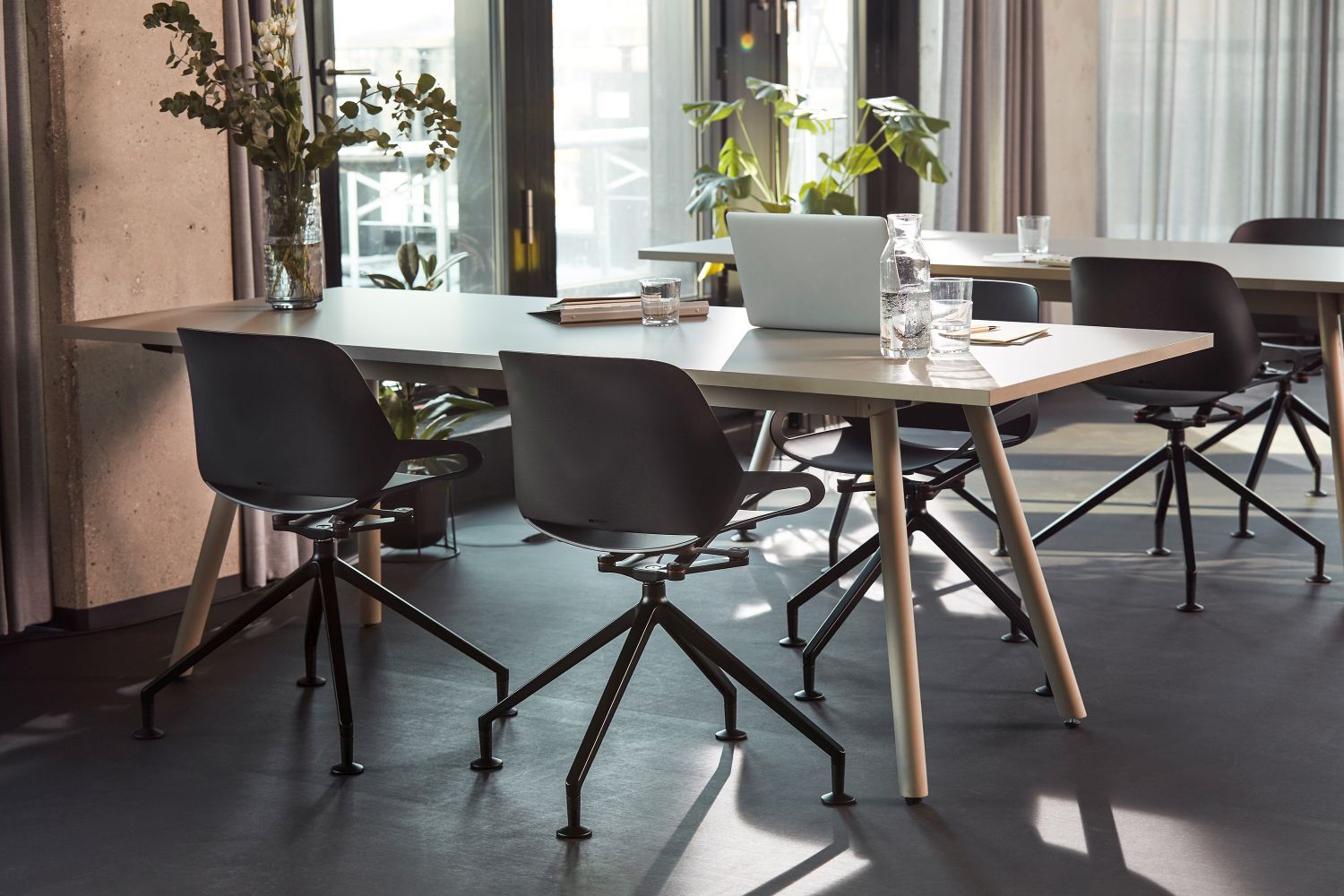 Moderner Besprechungstisch mit ergonomischen Stühlen in hellem Büroraum, Pflanzen im Hintergrund, Laptop und Wasserflasche.