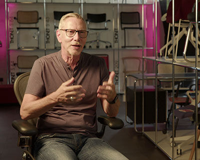 Mann in ergonomischem Stuhl sitzend, spricht vor einer Wand mit verschiedenen Stuhlmodellen im Hintergrund.