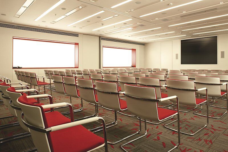 Leerer Konferenzraum mit rot-gepolsterten Stühlen, modernem Teppich und großem Bildschirm an der Wand.