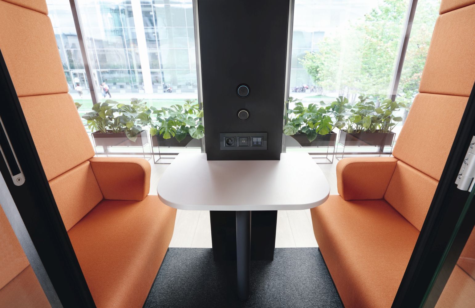 Zwei ergonomische Sessel mit Tisch in einer modernen Kabine, umgeben von großen Fenstern und grünen Pflanzen.