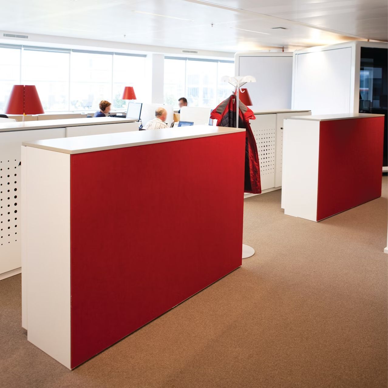 Helles Büro mit weißen Tischen, roten Frontpaneelen und Personen, die an Computern arbeiten.