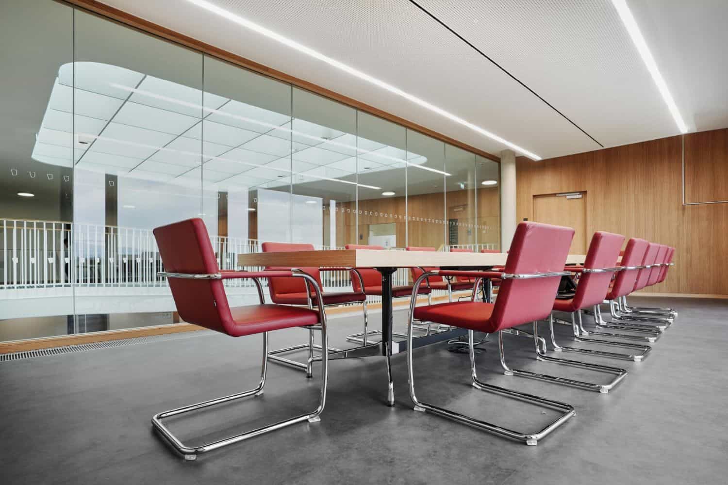 Moderner Konferenzraum mit roten, ergonomischen Stühlen und einem großen Tisch vor einer Glaswand.