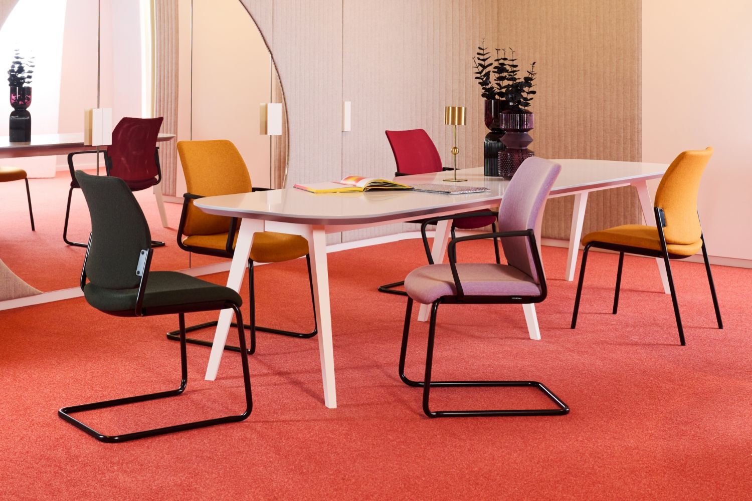 Moderner Besprechungsraum mit ergonomischen Stühlen in verschiedenen Farben um einen weißen Tisch auf rotem Teppich.