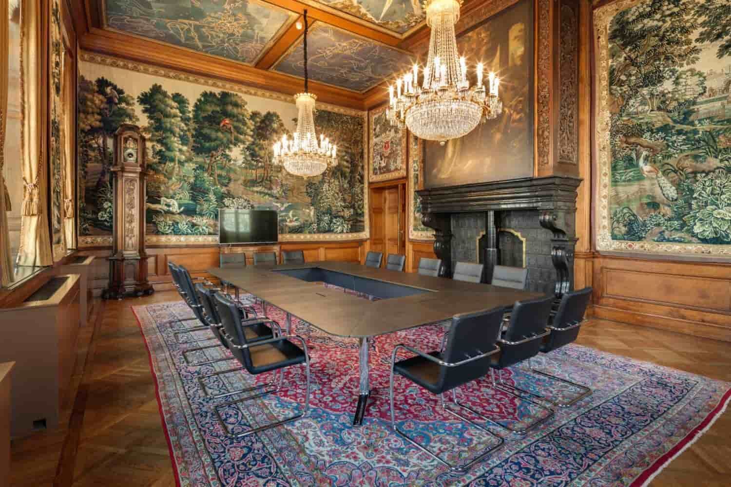Eleganter Konferenzraum mit großem Tisch, modernen Stühlen, antikem Kamin, Teppich und prunkvollen Kronleuchtern.