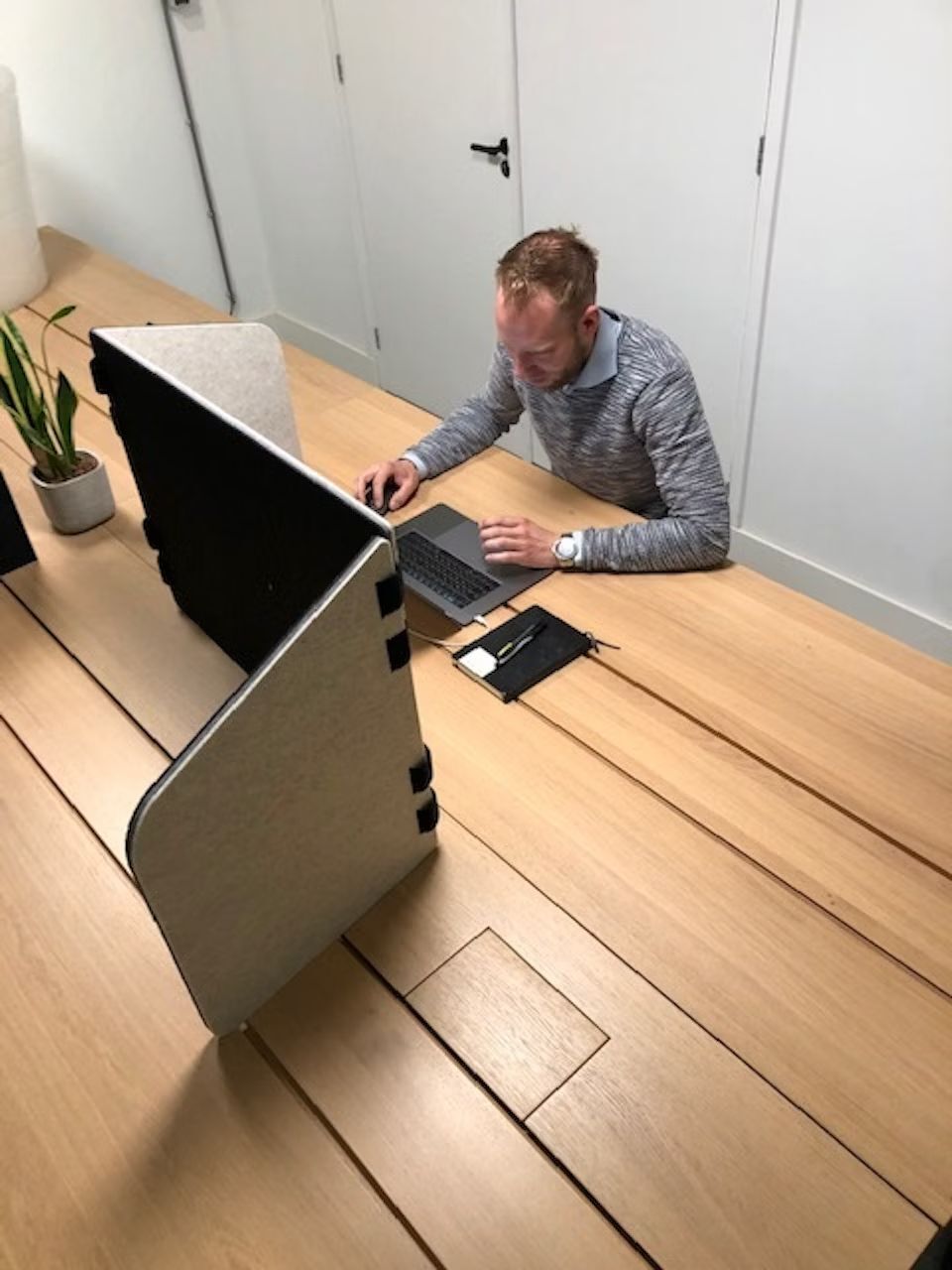 Person sitzt an einem Holztisch mit Laptop, umgeben von Trennwänden, in einem modernen Büro.