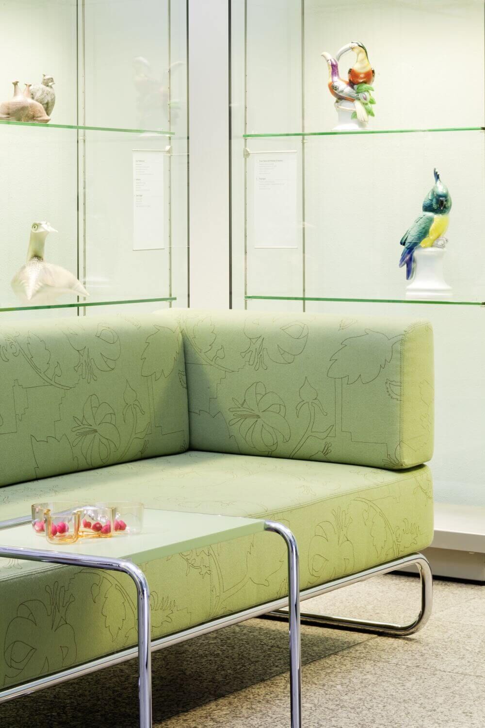 Grünes Sofa vor Glasvitrine mit dekorativen Vogelstatuen, betont modernes, ergonomisches Design für dynamisches Sitzen.