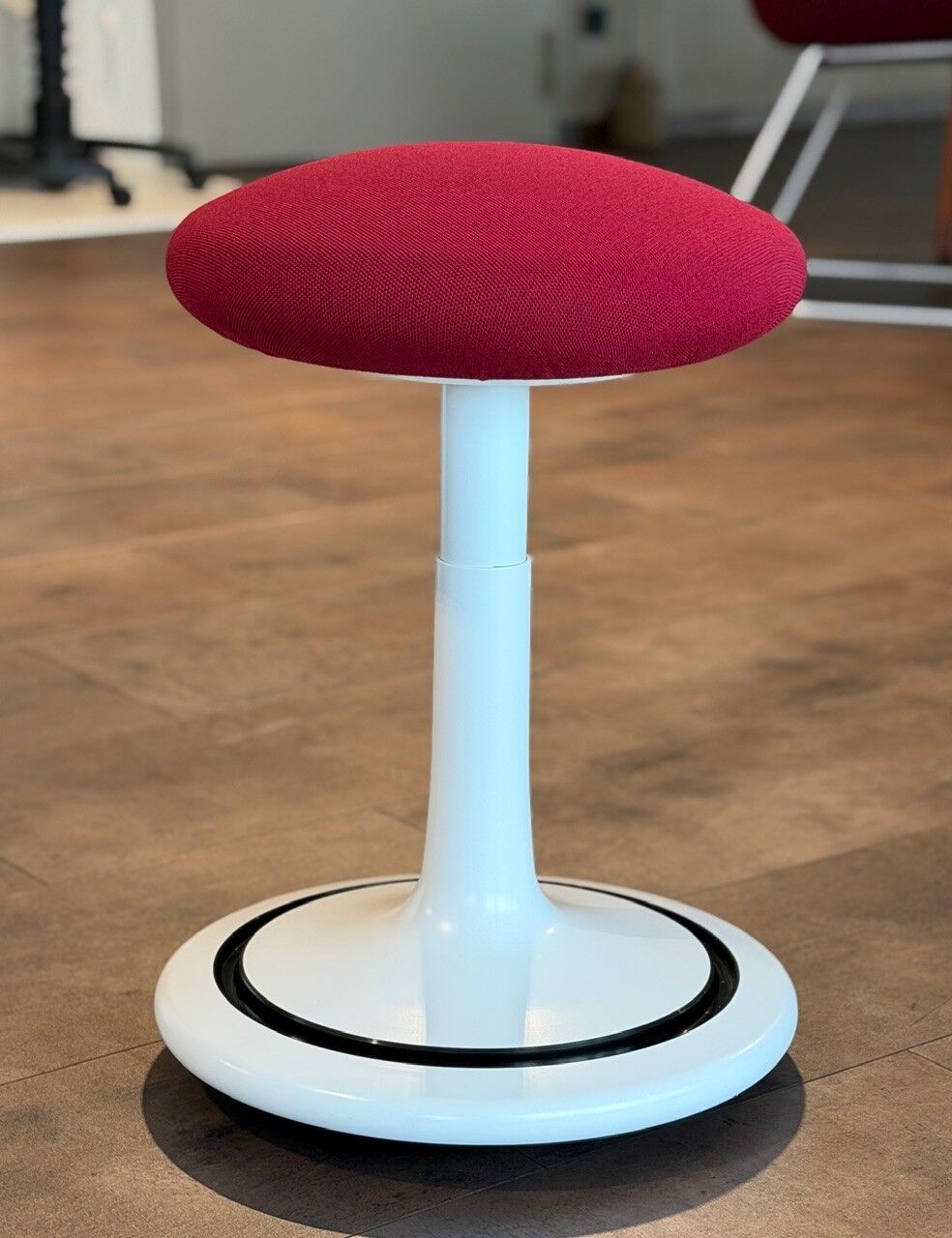 Ergonomischer Hocker mit rotem Polstersitz und weißem, runden Sockel auf Holzboden, ideal für bewegtes Sitzen.