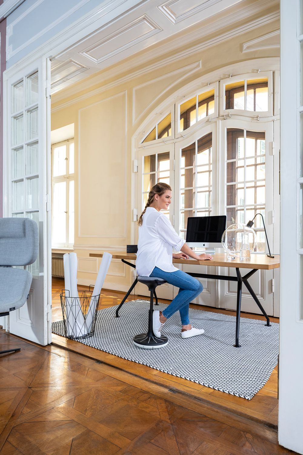 Frau sitzt auf ergonomischem Hocker an einem Schreibtisch in einem hellen, stilvollen Büro mit Holzboden.