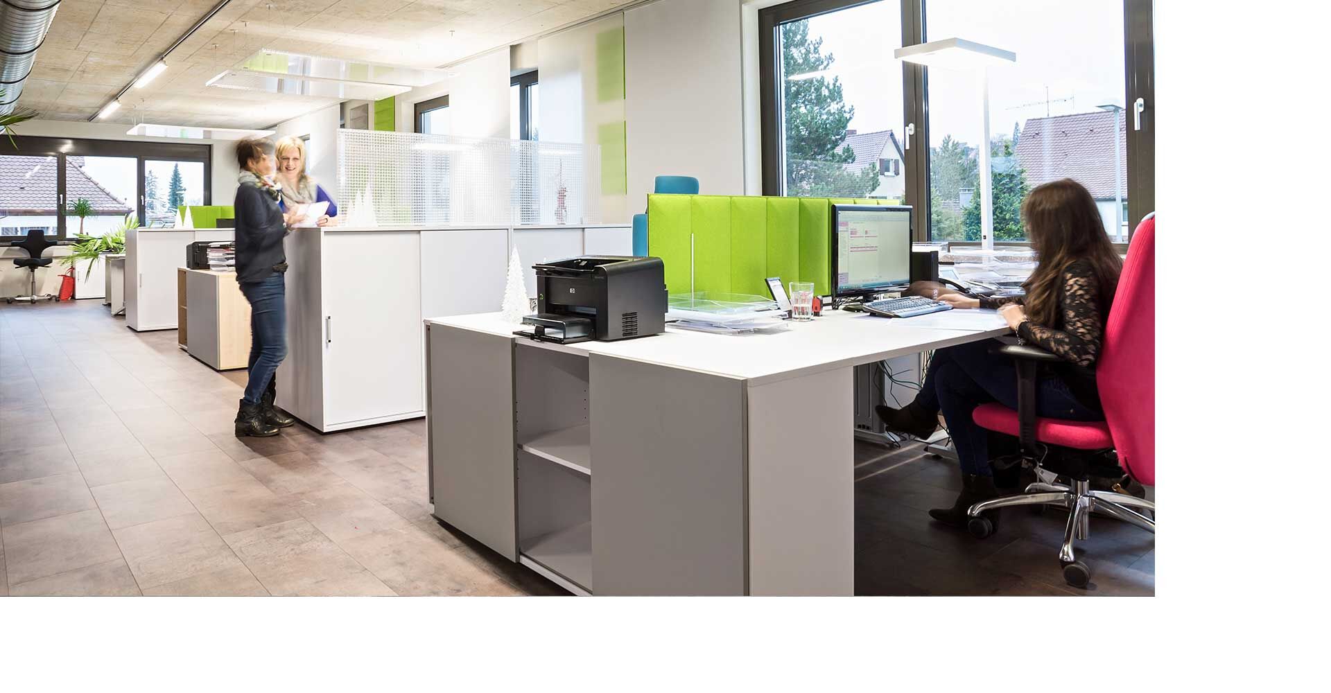 Modernes Büro mit ergonomischen Stühlen und Schreibtischen, zwei Frauen im Gespräch und eine arbeitet am Computer.