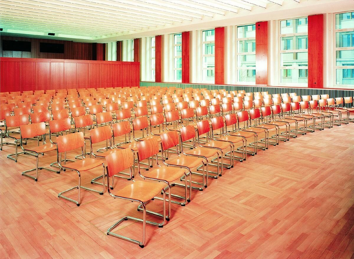 Großer Raum mit vielen leeren, hölzernen Konferenzstühlen auf hellem Parkettboden und großen Fensterflächen.