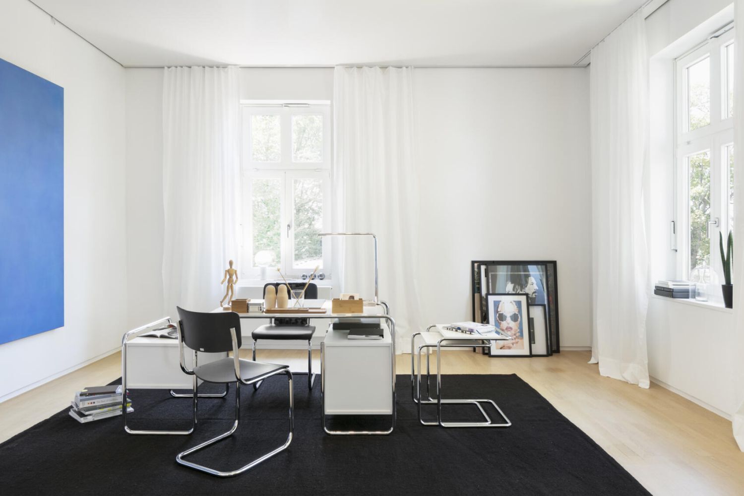 Modernes Büro mit ergonomischem Stuhl, minimalistischem Schreibtisch und großer Fensteransicht. Blaues Kunstwerk an der Wand.