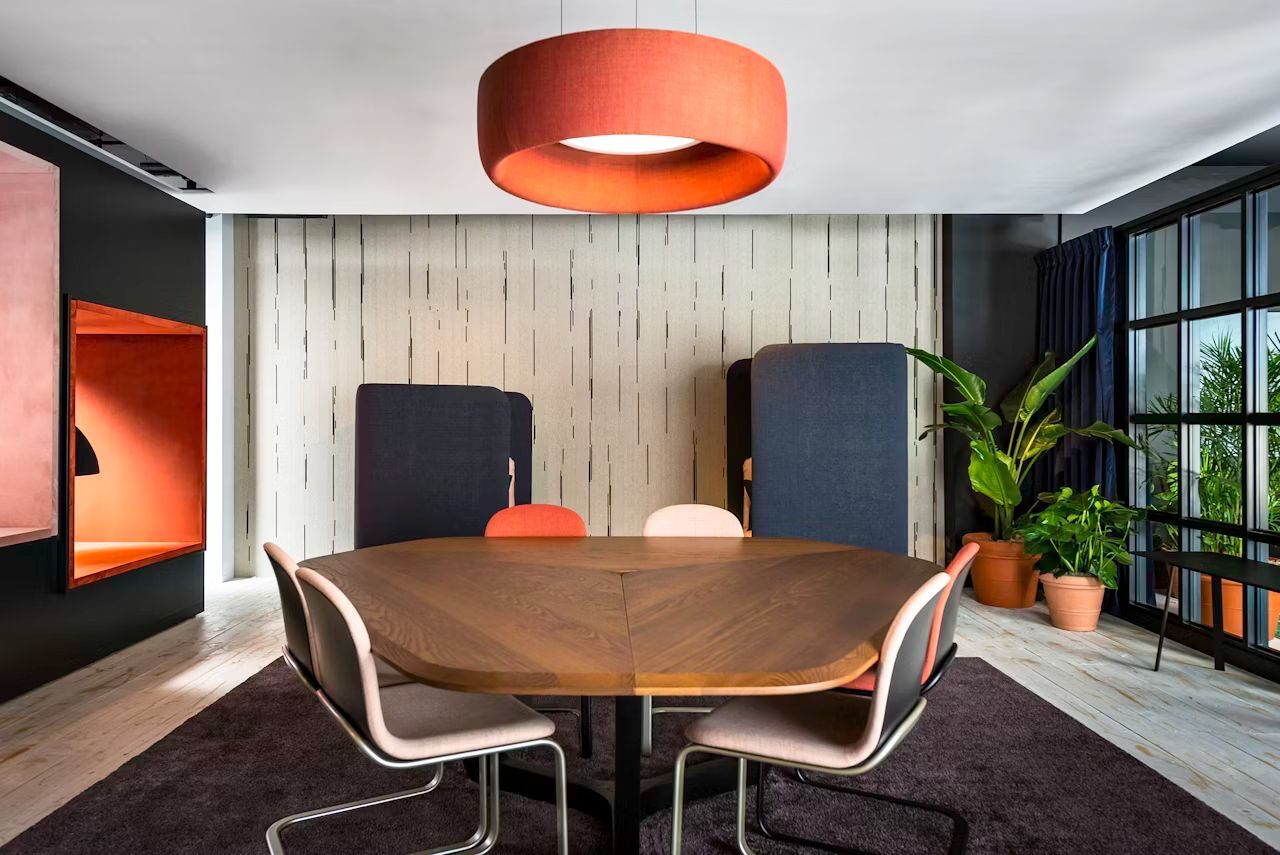 Moderner Besprechungsraum mit großem Holztisch, ergonomischen Stühlen und Pflanzen, beleuchtet durch eine orange Hängelampe.