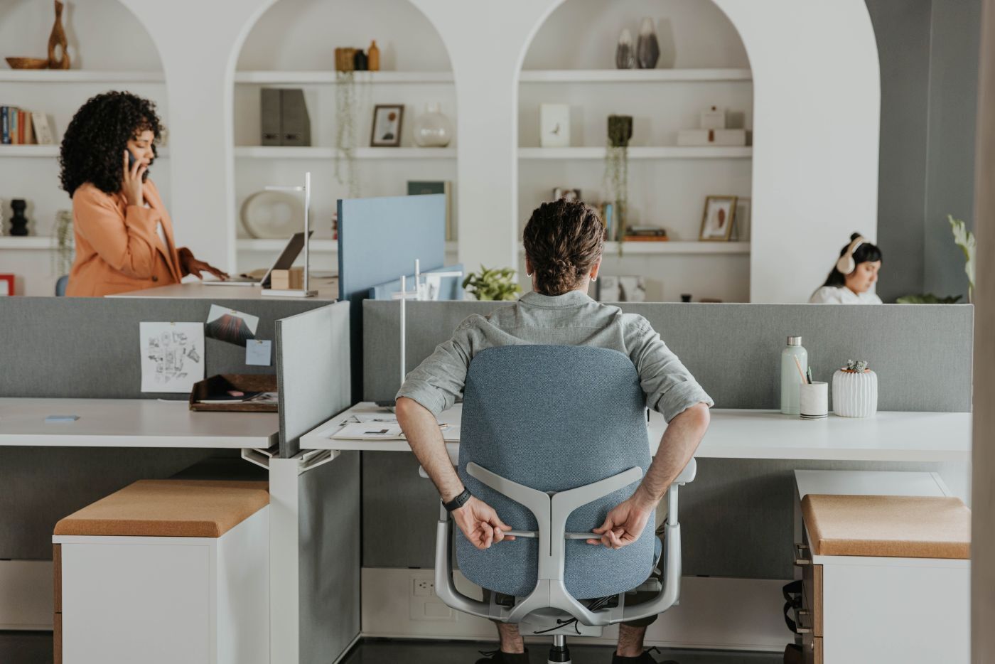 Person sitzt auf ergonomischem Bürostuhl, während andere Personen im modernen Büro arbeiten.