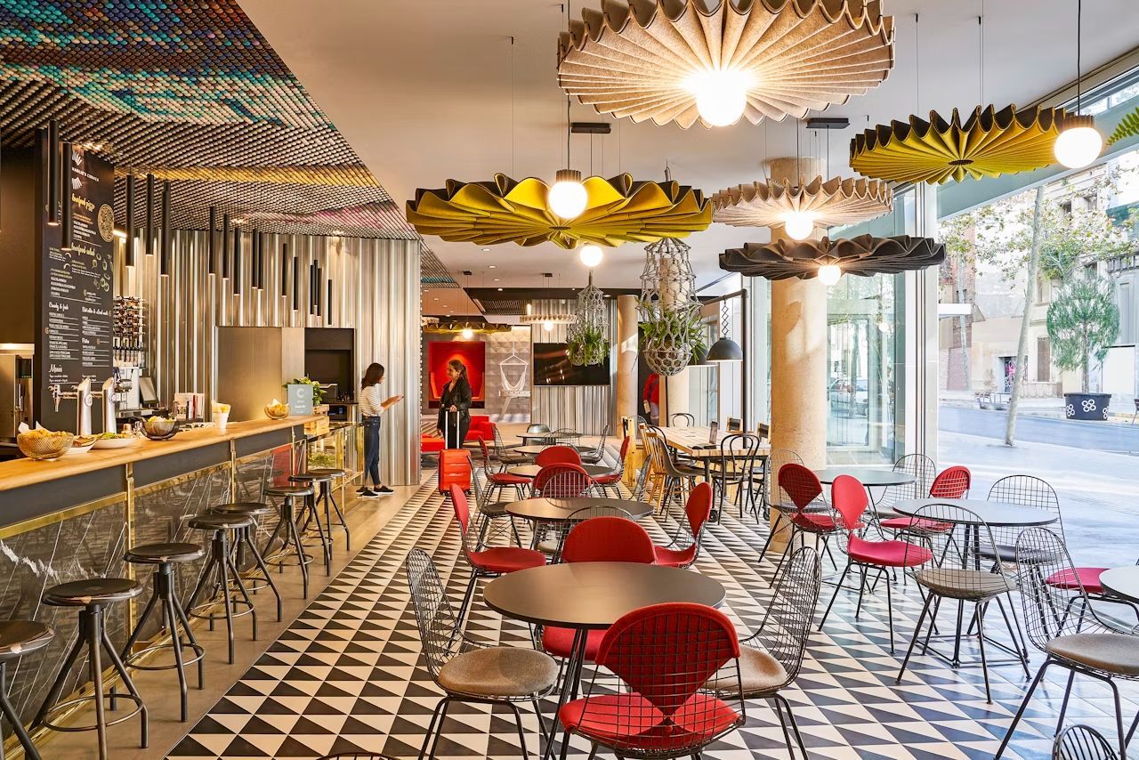 Modernes Café mit geometrischen Böden, bunten Möbeln und künstlerischen Deckenlampen. Zwei Personen im Gespräch an der Theke.