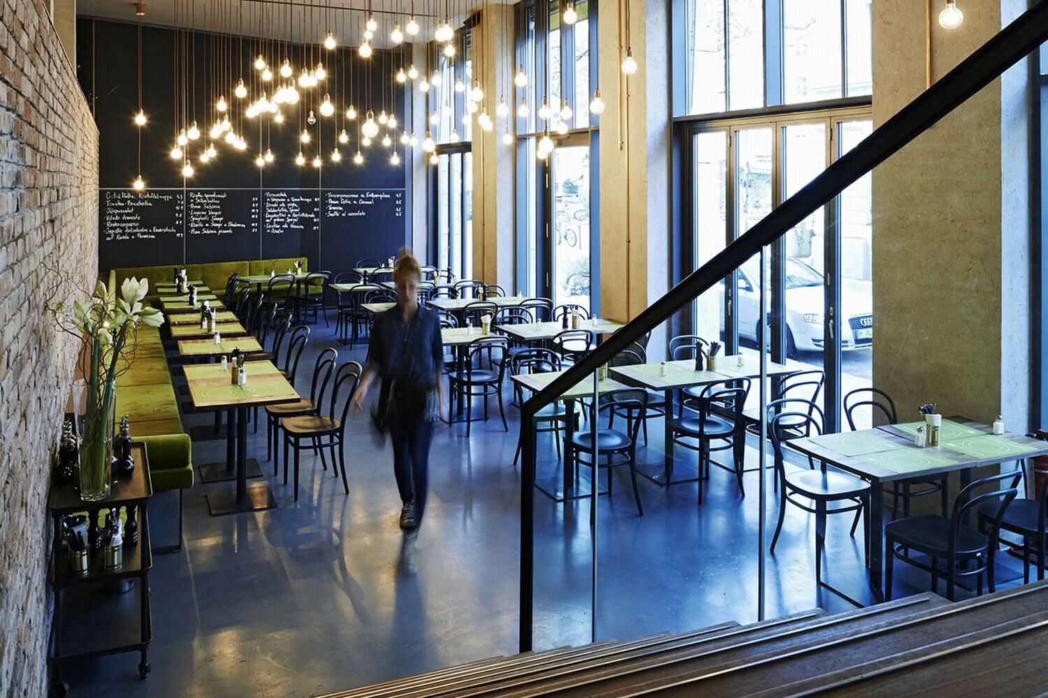 Modernes Café mit stylischer Beleuchtung, grünen Sitzbänken und großen Fenstern. Eine verschwommene Gestalt geht durch den Raum.