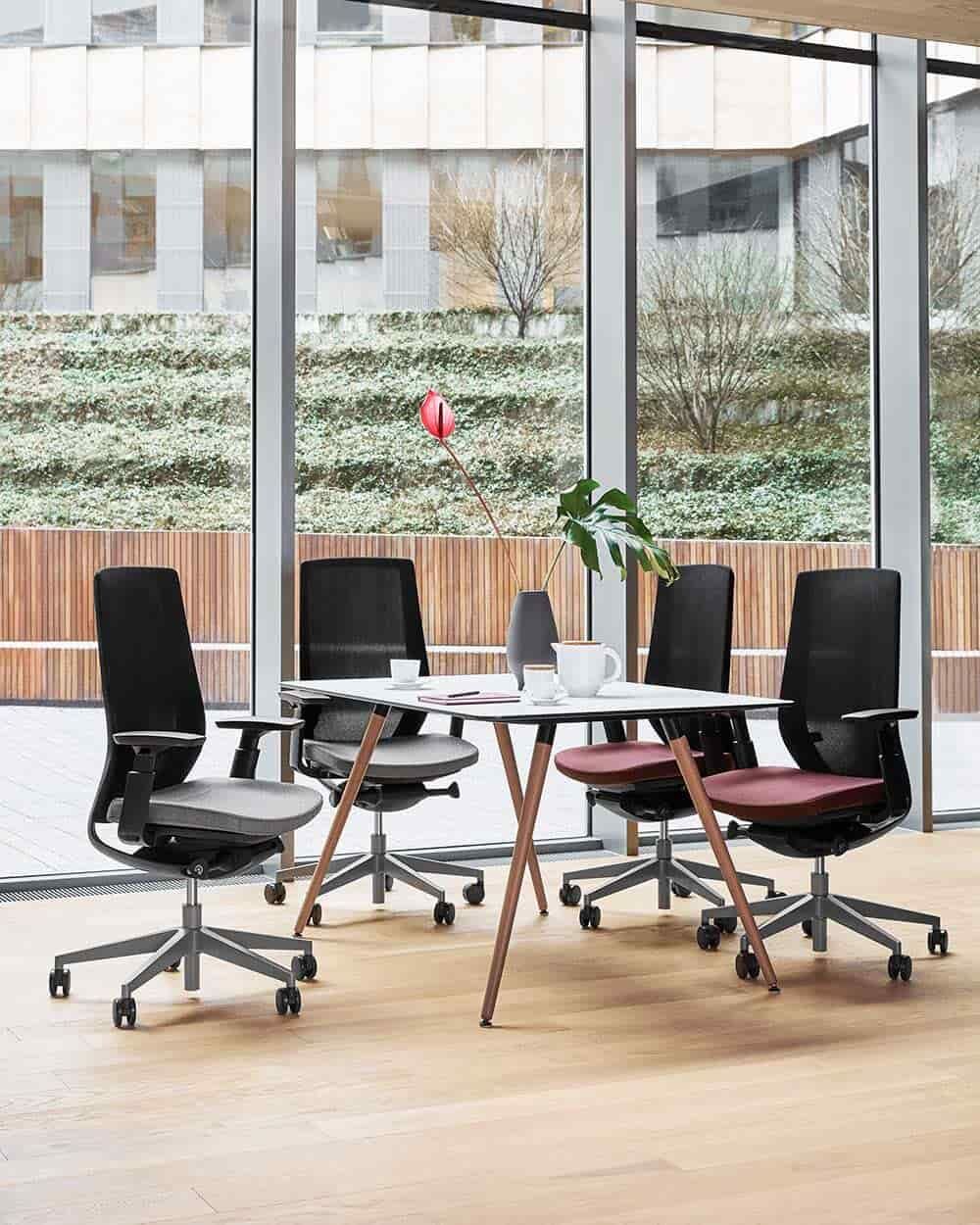 Ergonomische Bürostühle um einen minimalistischen Tisch, vor großen Glasfenstern mit Blick auf eine grüne Außenanlage.