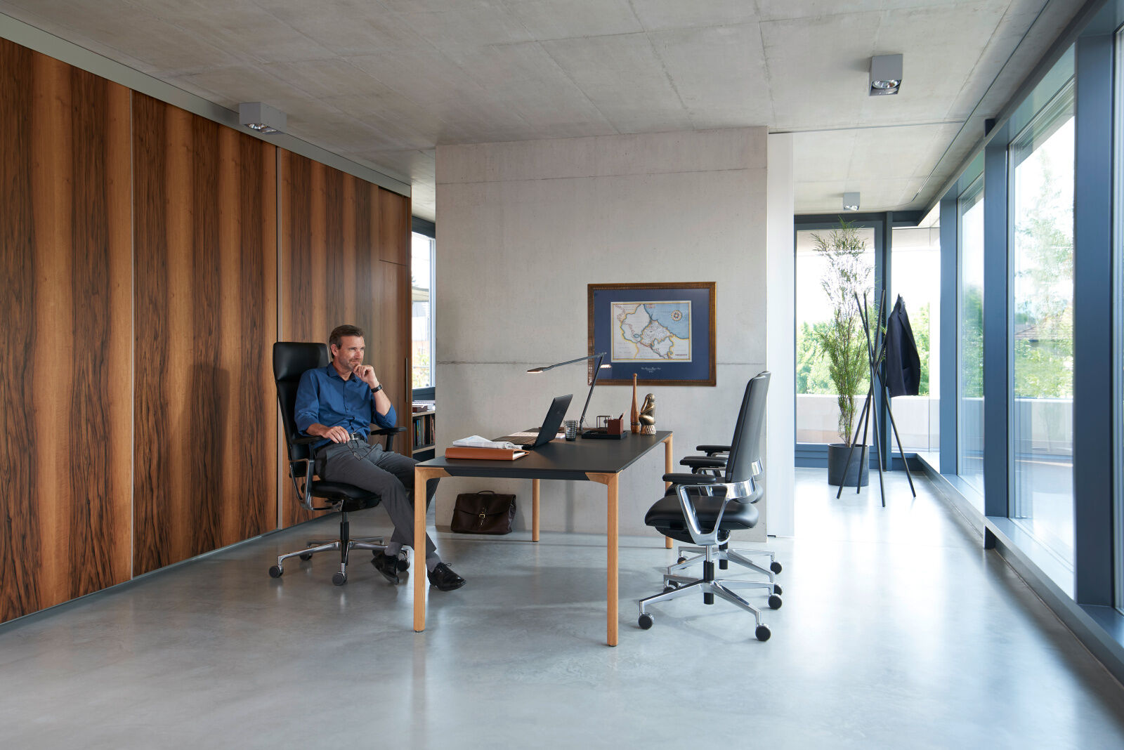 Mann sitzt auf ergonomischem Bürostuhl an modernem Schreibtisch in hell beleuchtetem Büro mit Holzwand und Fenster.
