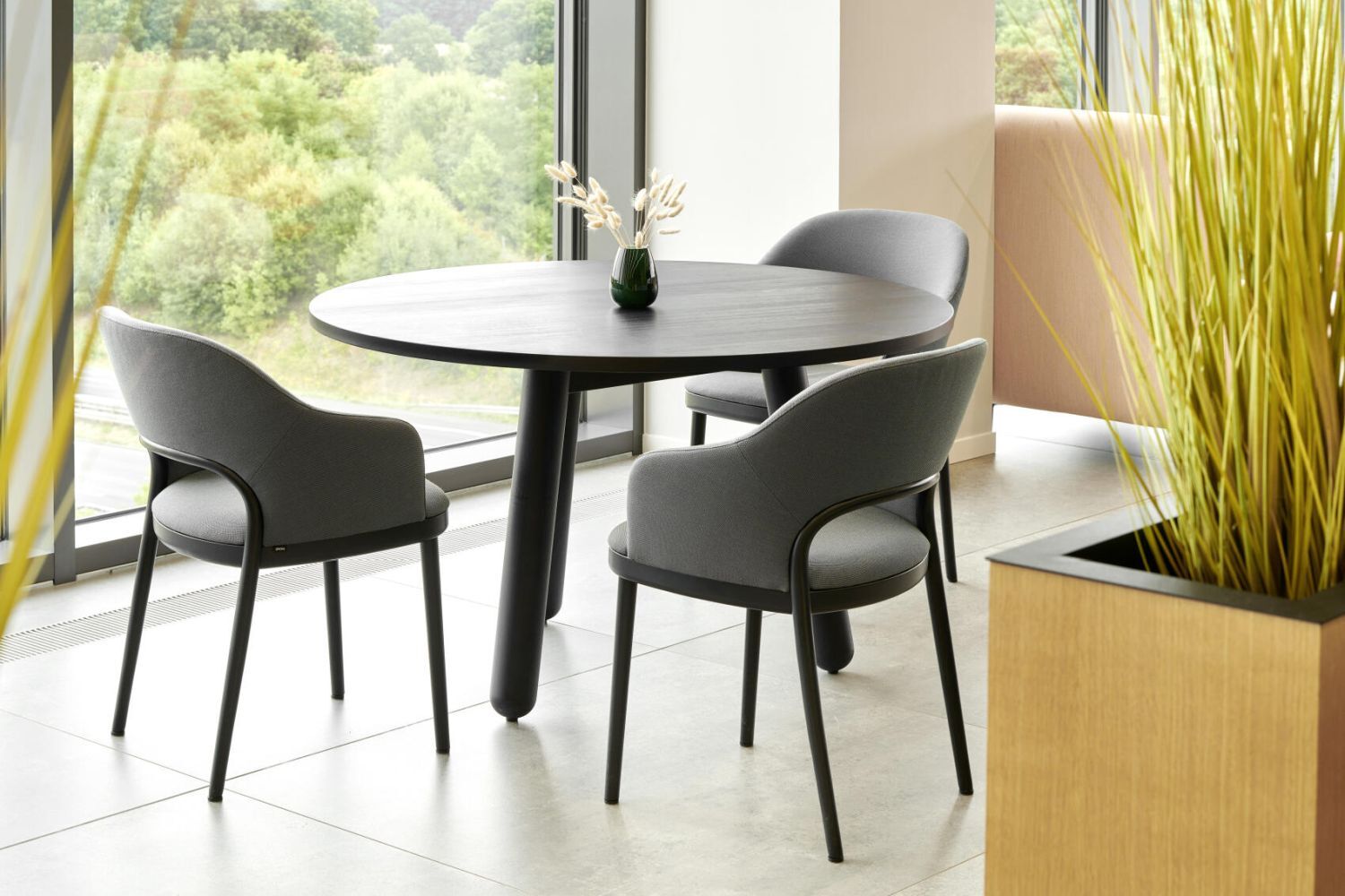 Runder Tisch mit drei grauen Stühlen in modernem, lichtdurchflutetem Raum mit großen Fenstern und Pflanzen.