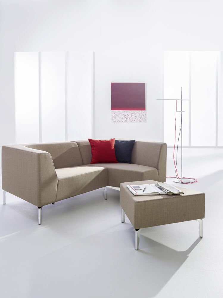 Minimalistisches Ecksofa mit roten Kissen und passendem Hocker in hellem Raum mit moderner Kunst.