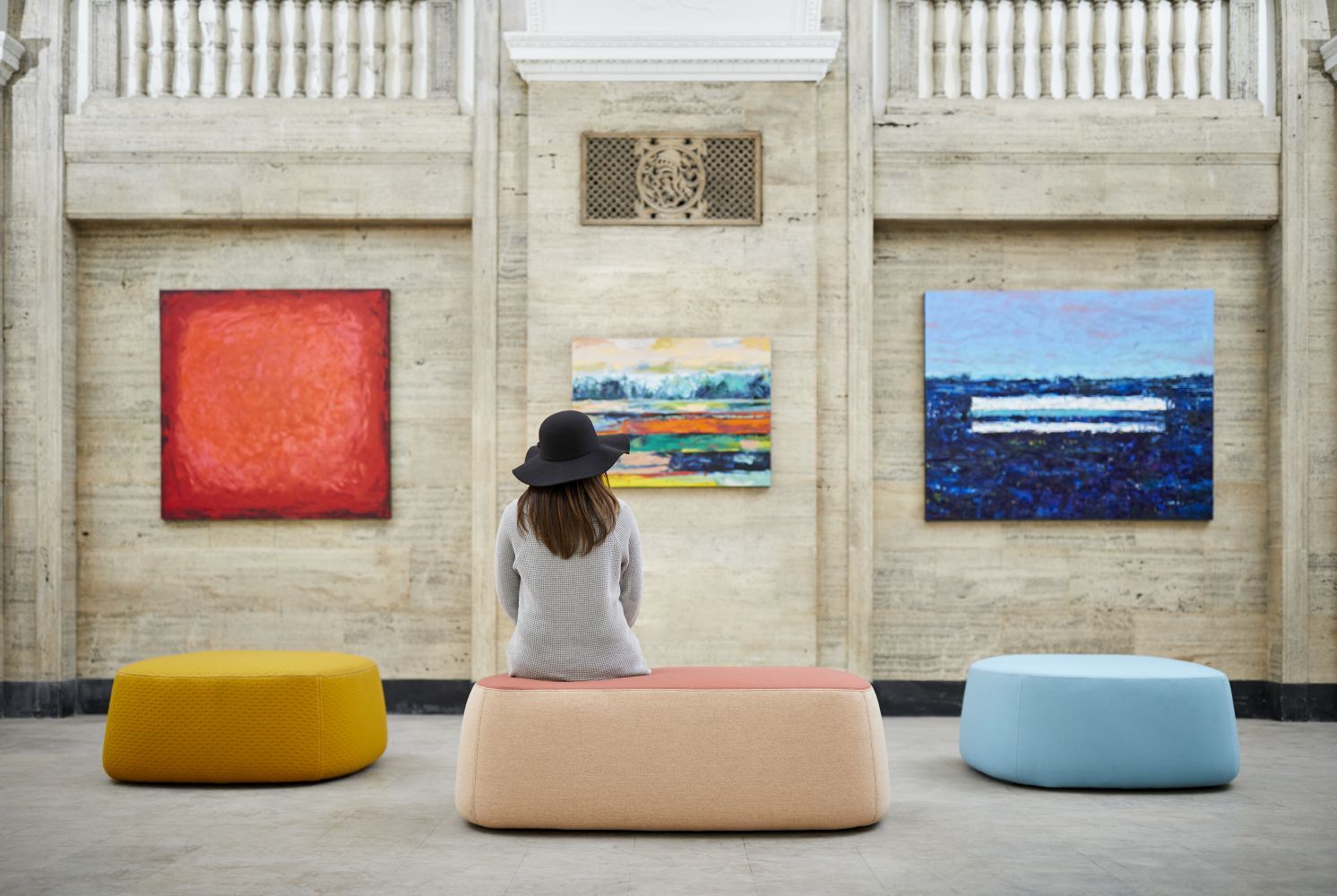 Frau mit Hut sitzt auf einem Polsterhocker in moderner Kunstgalerie mit bunten Gemälden an der Wand.