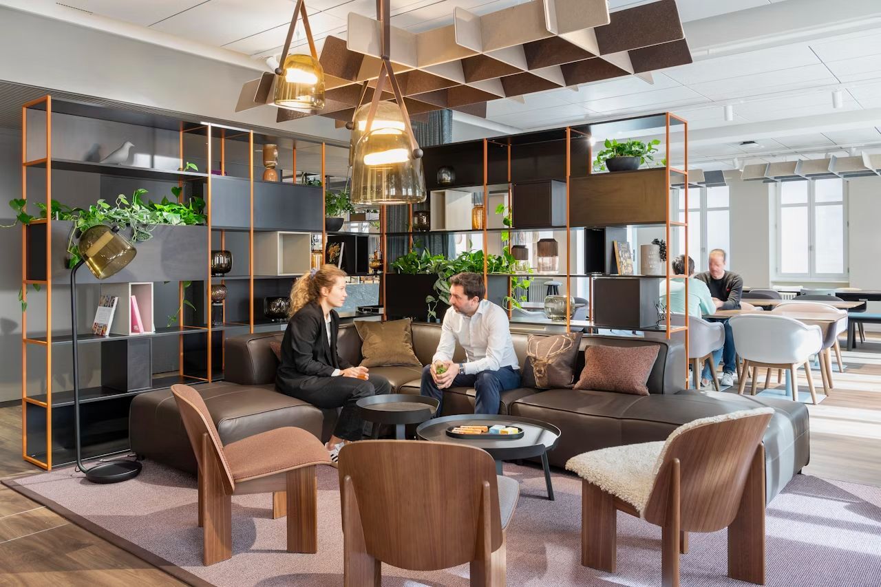 Moderne Lounge-Ecke mit Pflanzen, Regalen und zwei Personen, die auf einem Ledersofa sprechen.