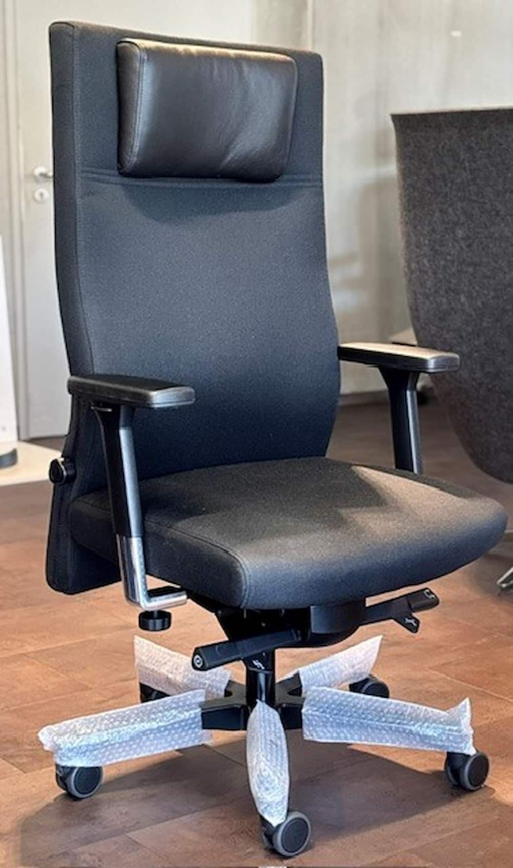 Ergonomischer Bürostuhl mit hoher Rückenlehne und Kopfstütze, auf Holzboden, mit Folie umwickelte Stuhlbeine.