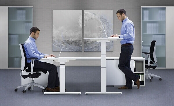 Zwei Männer an höhenverstellbaren Schreibtischen, einer sitzend und einer stehend, in einem modernen Büro.