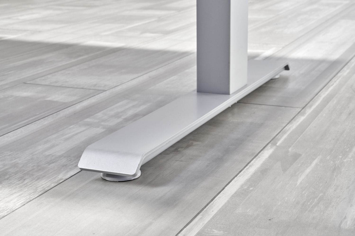 Nahaufnahme des stabilen, weißen Metallfußes eines ergonomischen Tischgestells auf hellgrauem Holzboden.