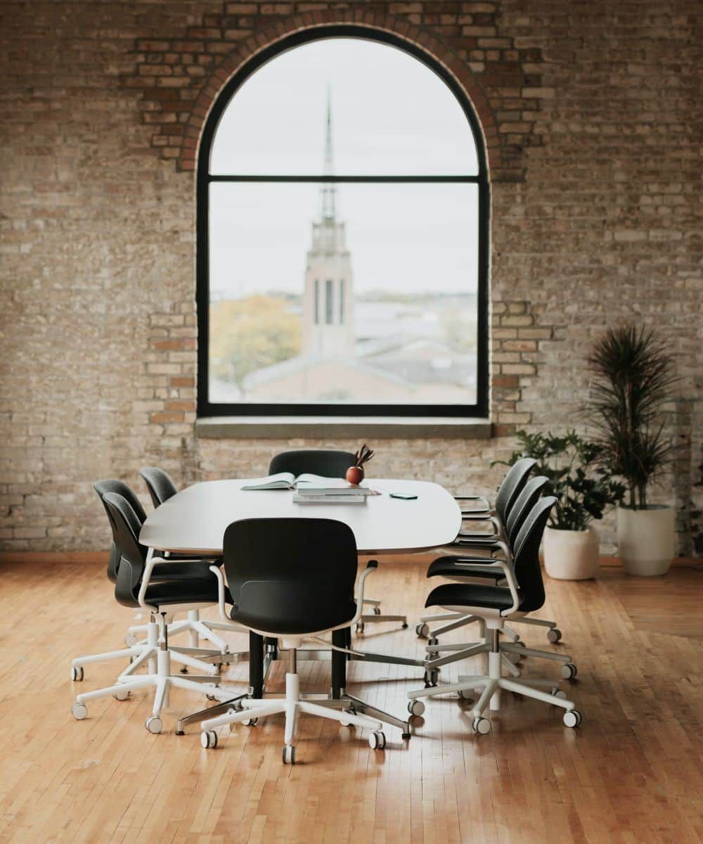 Konferenzraum mit ergonomischen Stühlen um einen Tisch, großes Fenster, freiliegende Ziegelwand im Hintergrund.