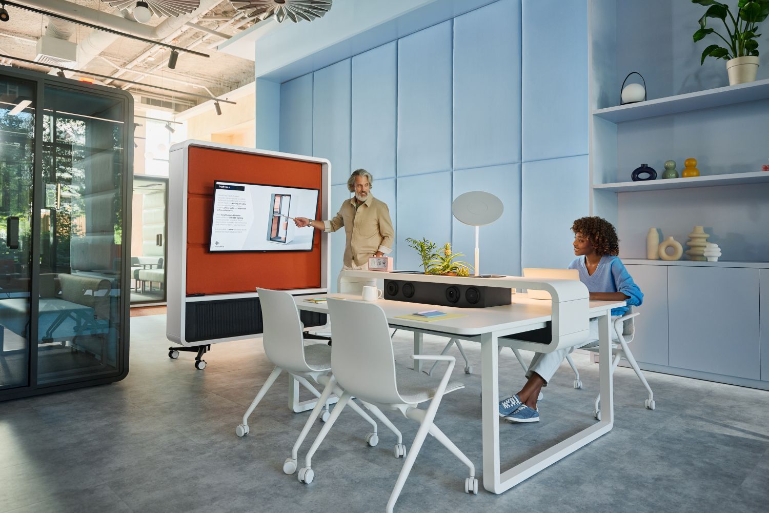 Person präsentiert an einem Bildschirm im modernen Büro. Ergonomische Stühle und Tisch im Vordergrund.