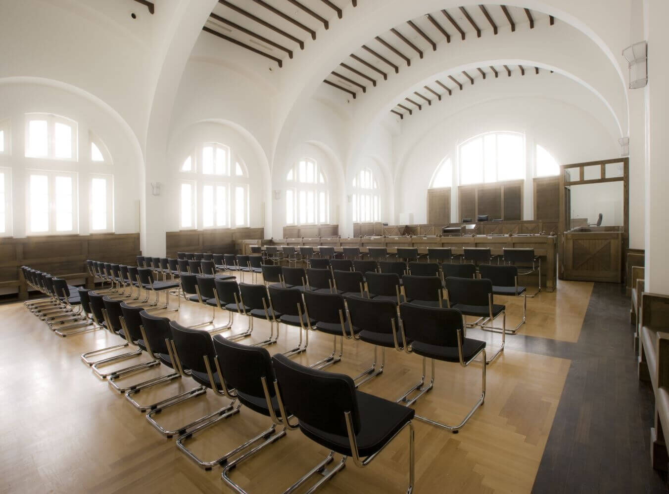Großer, heller Konferenzsaal mit Reihen von schwarzen Stühlen auf einem Parkettboden und hoher Decke.