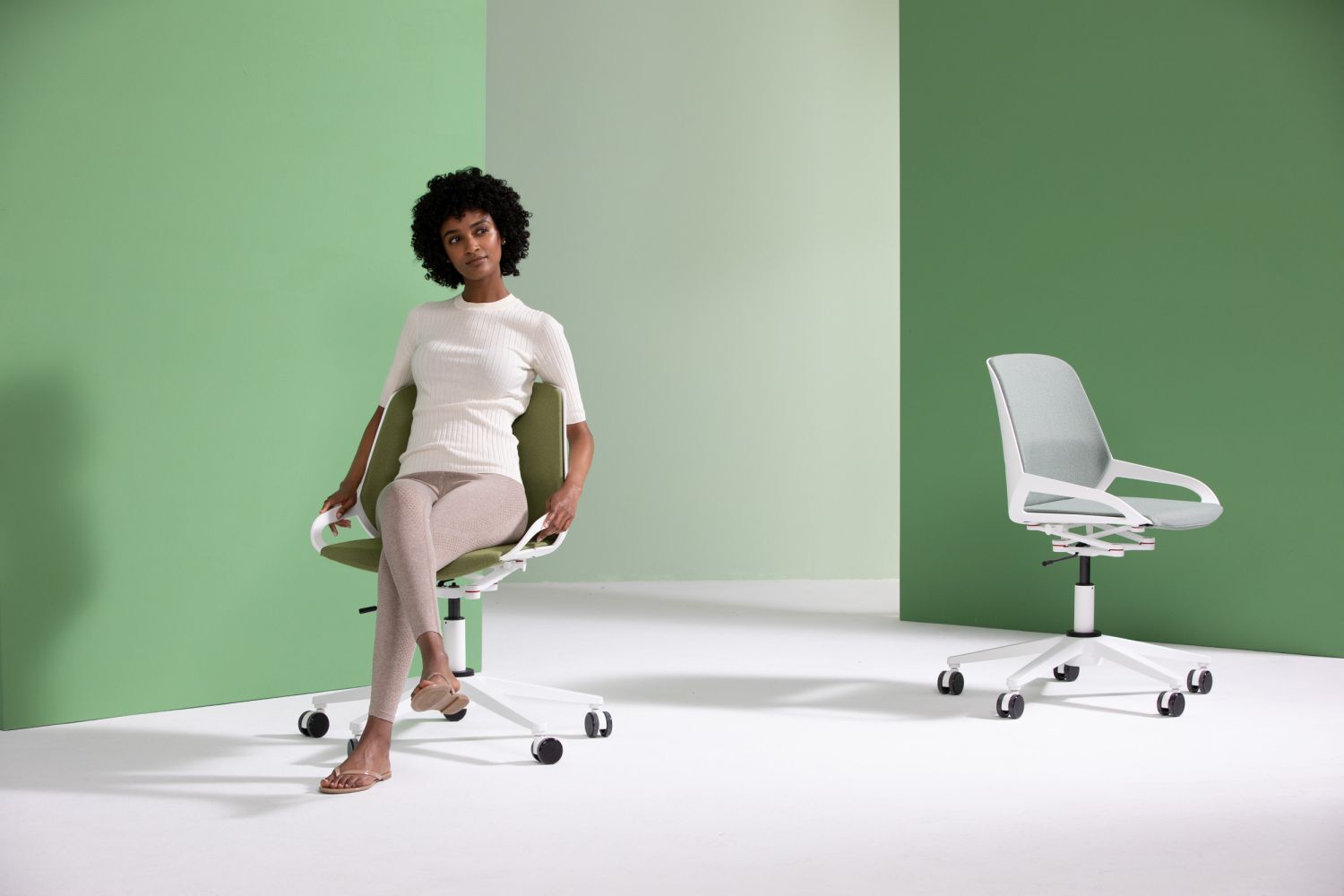 Frau sitzt auf einem ergonomischen Bürostuhl vor grüner Kulisse, neben einem zweiten, unbesetzten Stuhl.