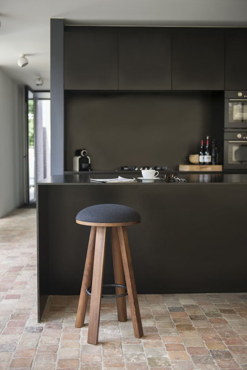 Moderner Holzbarhocker mit gepolstertem Sitz in minimalistischer Küche mit Fliesenboden und dunklen Schränken.