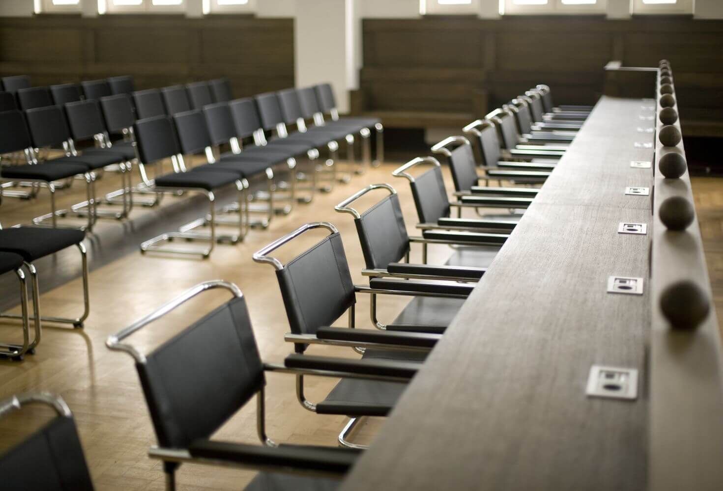 Reihen schwarzer Stühle und Tische in einem Konferenzraum mit Holzfußboden und Fenstern im Hintergrund.