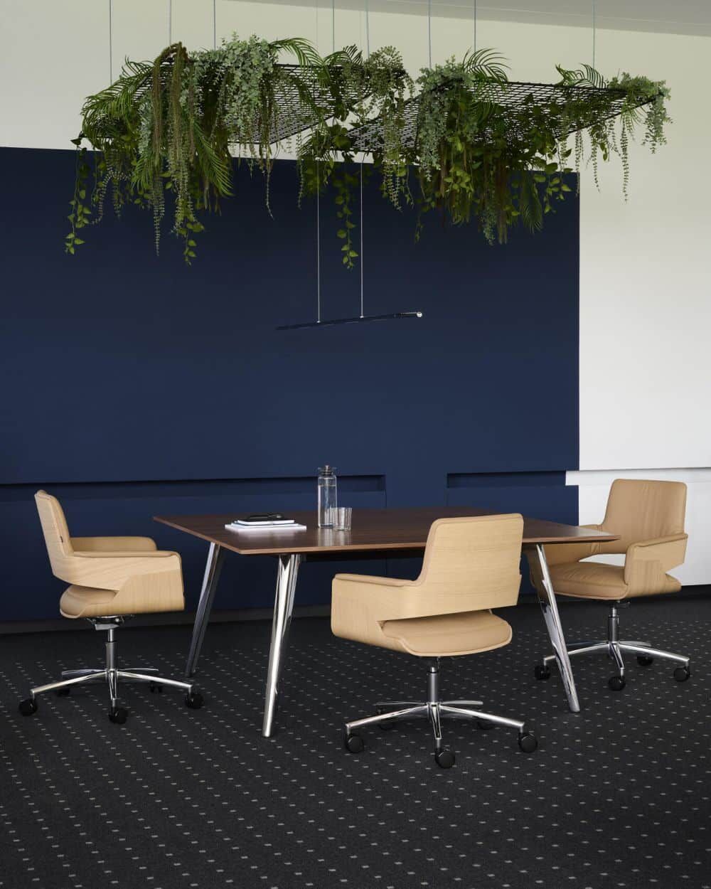 Ergonomische Bürostühle an einem Holztisch, hängende Pflanzen darüber, vor einer dunkelblauen Wand.