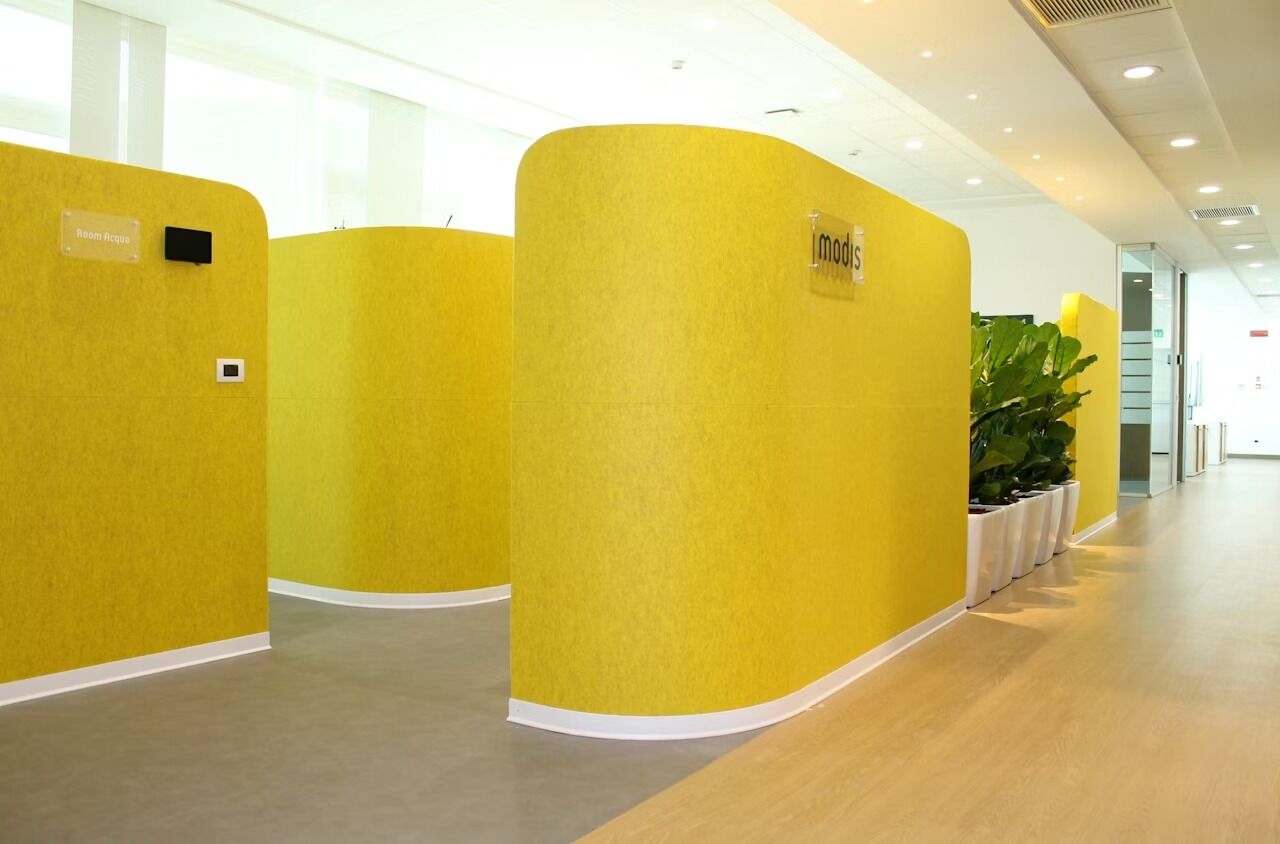 Modernes Büro mit gelben, gewölbten Trennwänden, natürlichem Licht und einer Reihe großer Pflanzen.