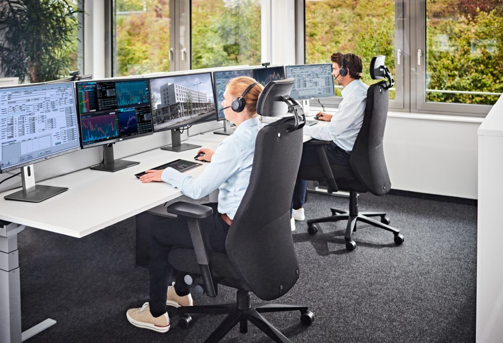 Zwei Personen arbeiten an Schreibtischen mit ergonomischen Stühlen und mehreren Bildschirmen in einem modernen Büro.