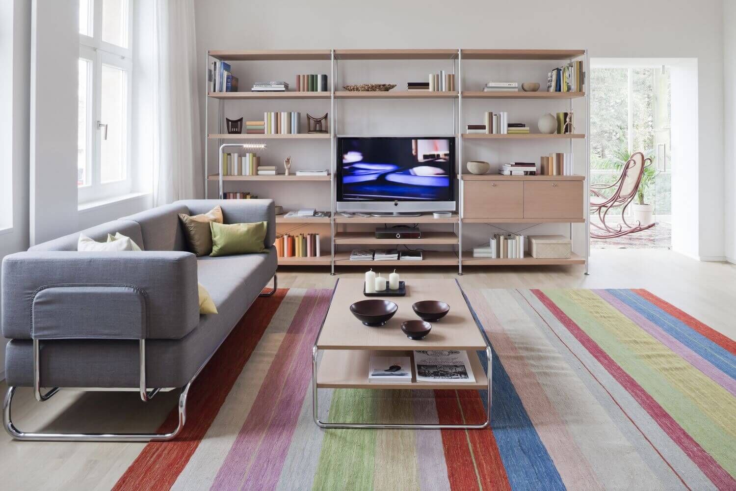 Helles Wohnzimmer mit grauem Sofa, Regalen, Fernseher und farbig gestreiftem Teppich auf Holzboden.