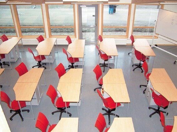 Klassenzimmer mit hellen Holztischen und roten ergonomischen Bürostühlen, ausgerichtet in mehreren Reihen vor großen Fenstern.