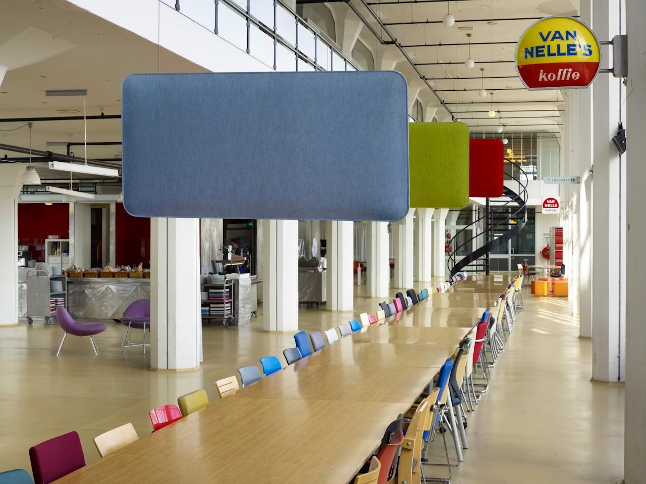 Langer Tisch mit bunten Stühlen in einem hellen, modernen Loft mit akustischen Deckensegeln und Wendeltreppe.