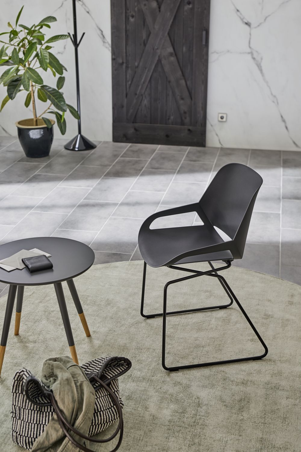 Moderner schwarzer Stuhl auf Teppich, neben Tisch mit Geldbörse und Topfplanze vor Holztür.