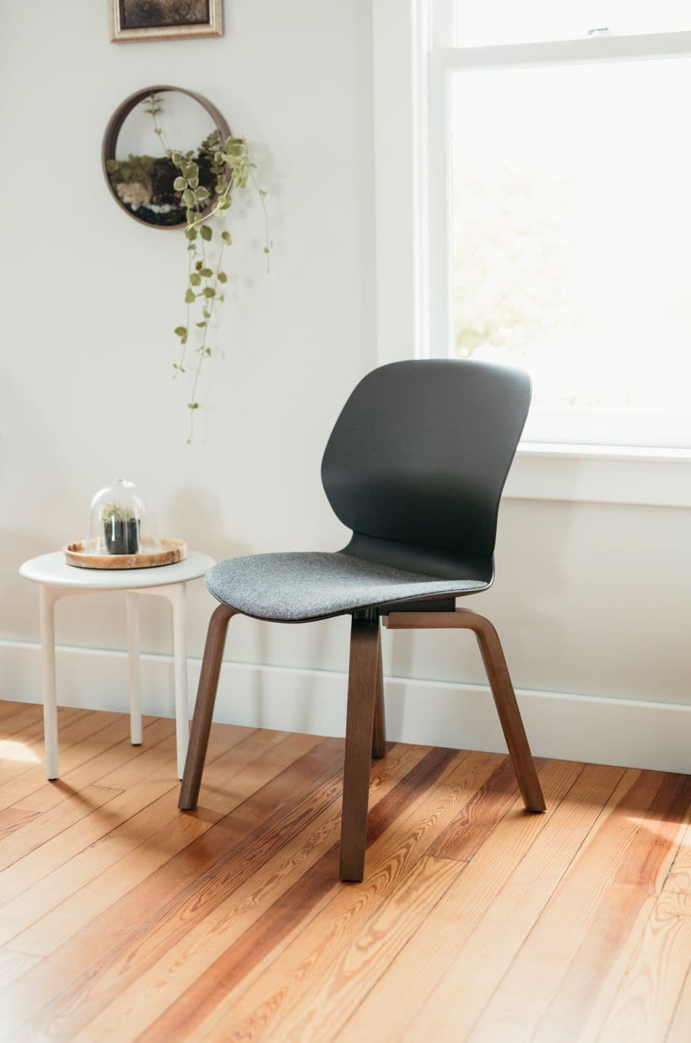 Ergonomischer Stuhl mit Holzbasis und grauem Sitzpolster, neben einem kleinen weißen Tisch in hellem Raum.