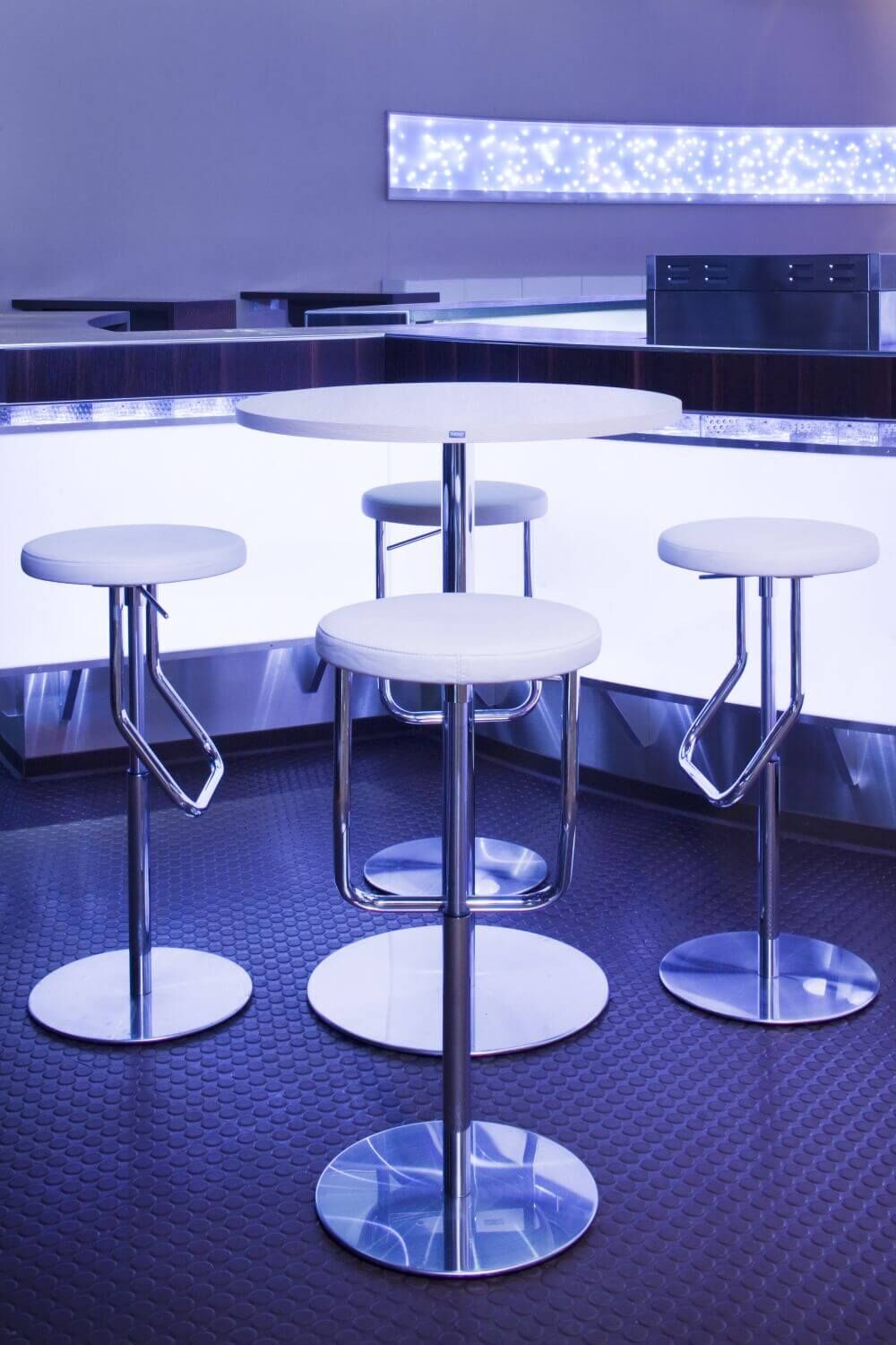 Vier moderne Barhocker vor beleuchtetem Tresen im eleganten, futuristischen Design mit Metallakzenten.